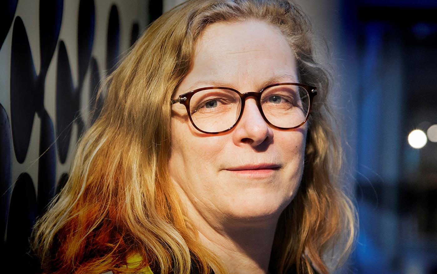 Kvinna med lockigt hår och glasögon tittar mot kameran