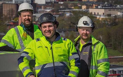 Tre män stående på hög höjd ler mot kameran i sina arbetskläder och hjälmar på huvudet